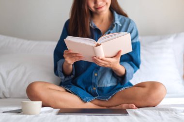 Evdeki rahat beyaz bir yatakta oturmuş kitap okuyan güzel bir genç kadın.