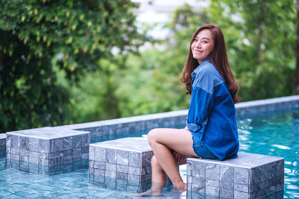 Portrait image of a beautiful young asian woman enjoyed sitting by swimming pool with green nature background