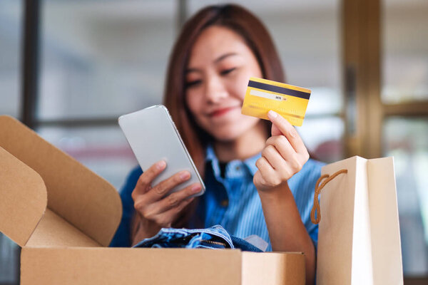 Closeup image of a beautiful young woman using mobile phone and credit card for online shopping with shopping bag and postal parcel box of clothing on the table