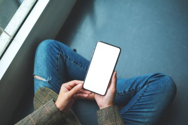 Zeminde otururken elinde boş beyaz masaüstü ekran olan cep telefonu tutan bir kadının en üst görünüm görüntüsü