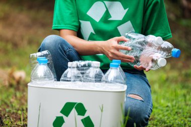 Açık havada plastik şişe toplayıp geri dönüşüm kutusuna koyan bir kadın.