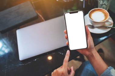 Masasında dizüstü bilgisayarı olan, boş beyaz masaüstü ekranlı cep telefonu tutan bir kadının görüntüsü.