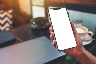 Masasında dizüstü bilgisayarı olan, boş beyaz masaüstü ekranlı cep telefonu tutan bir kadının görüntüsü.