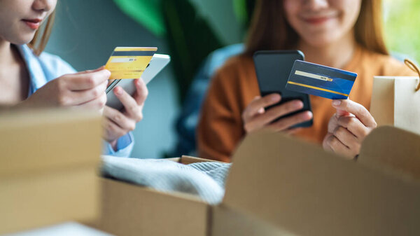 Two women using mobile phone and credit card for online shopping with shopping bag and postal parcel box of clothing on the table