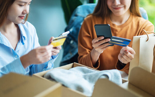 Two women using mobile phone and credit card for online shopping with shopping bag and postal parcel box of clothing on the table