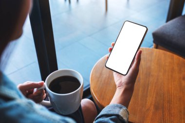 Kahve içerken elinde boş beyaz masaüstü ekranıyla cep telefonu tutan bir kadının görüntüsü