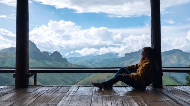 Dişi bir gezgin oturmuş, güzel dağlara bakıyor ve ahşap balkonda doğa manzarasına bakıyor.