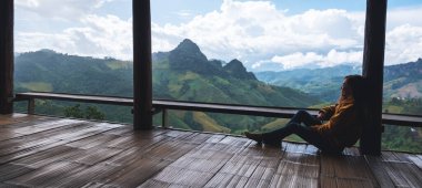 Dişi bir gezgin oturmuş, güzel dağlara bakıyor ve ahşap balkonda doğa manzarasına bakıyor.