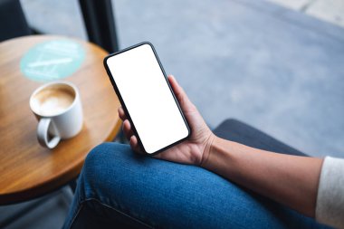 Masasında kahve fincanı olan boş beyaz masaüstü ekranlı cep telefonu tutan bir kadının görüntüsü.