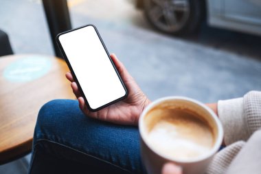 Kahve içerken elinde boş beyaz masaüstü ekranıyla cep telefonu tutan bir kadının görüntüsü 