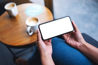 Masasında kahve fincanı olan boş beyaz masaüstü ekranlı cep telefonu tutan bir kadının görüntüsü.