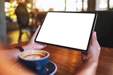 Elinde tablet olan, boş beyaz masaüstü ekranı olan, ahşap masada kahve fincanı olan bir kadının görüntüsü.