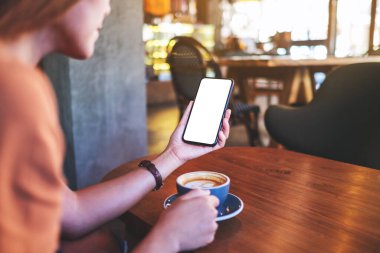 Kahve içerken elinde boş beyaz masaüstü ekranıyla cep telefonu tutan bir kadının görüntüsü 
