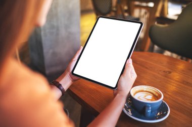 Elinde tablet olan, boş beyaz masaüstü ekranı olan, ahşap masada kahve fincanı olan bir kadının görüntüsü.