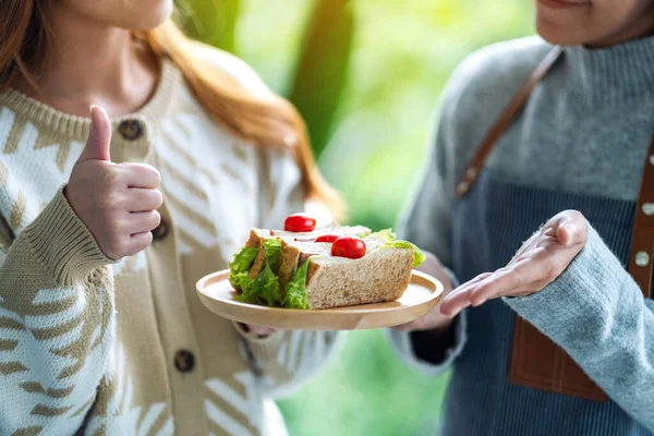 Bir garsonun tahtadan tabakta tam buğday sandviçi servis ederken, bir müşterinin el işaretini kapatırken görüntüsü.