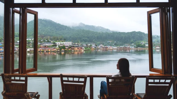 Genç bir kadın balkonda oturuyor ve dağ köyünde güzel bir göle bakıyor.