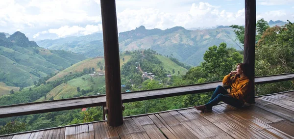 Dişi bir gezgin oturmuş, güzel dağlara bakıyor ve ahşap balkonda doğa manzarasına bakıyor.