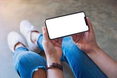 Yerde otururken elinde beyaz ekranlı siyah bir cep telefonu tutan bir kadının görüntüsü.