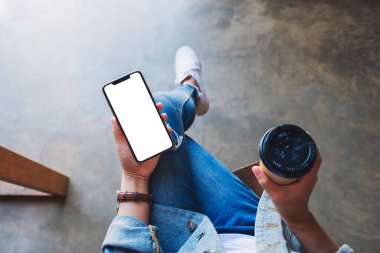 Üst görünümde, elinde siyah bir cep telefonu olan bir kadının kahve fincanı olan beyaz ekranlı görüntüsü var.