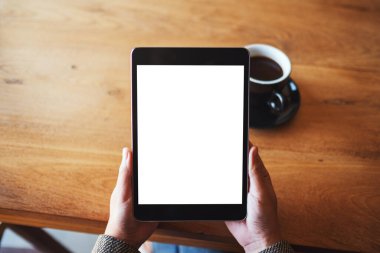 Top view mockup image of hands holding black tablet pc with blank white screen with coffee cup on wooden table 