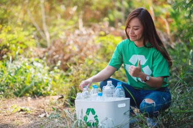 An asian woman making thumbs up hand sign while collecting garbage plastic bottles into a recycle bin in the outdoors