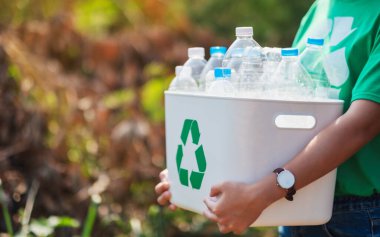 A woman collecting garbage and holding a recycle bin with plastic bottles in the outdoors