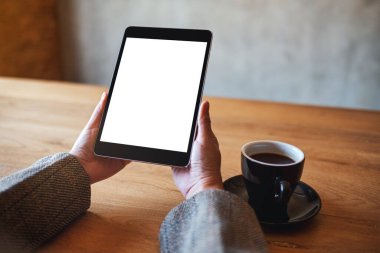 Siyah tabletli, beyaz ekranlı ve ahşap masada kahve fincanı olan ellerin görüntüsü. 