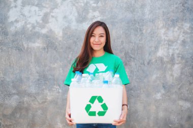 Elinde plastik şişelerle geri dönüşüm kutusunu tutan Asyalı bir kadın beton duvarın önünde duruyor.