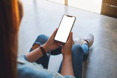 Yerde otururken elinde beyaz ekranlı siyah bir cep telefonu tutan bir kadının görüntüsü.