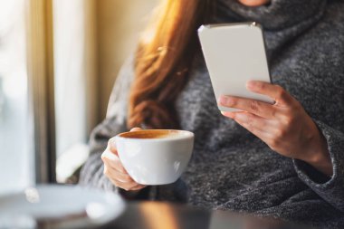 Kafede kahve içerken cep telefonu tutan ve kullanan bir kadının yakın plan fotoğrafı.