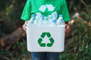 A woman collecting garbage and holding a recycle bin with plastic bottles in the outdoors