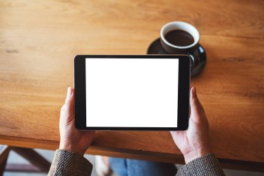 Top view mockup image of hands holding black tablet pc with blank white screen with coffee cup on wooden table 