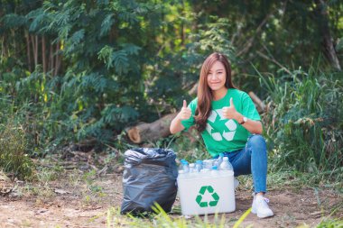 An asian woman making thumbs up hand sign while collecting garbage plastic bottles into a recycle bin in the outdoors