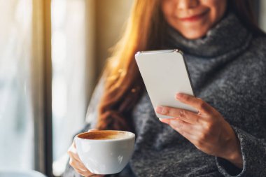 Kafede kahve içerken cep telefonu tutan ve kullanan bir kadının yakın plan fotoğrafı.