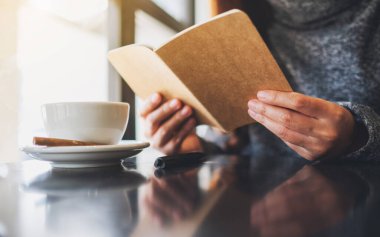 Closeup image of a woman holding and reading a book with coffee cup on the table