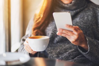 Kafede kahve içerken cep telefonu tutan ve kullanan bir kadının yakın plan fotoğrafı.