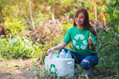 Güzel bir Asyalı kadın, dışarıda çöp kutusuna plastik şişe toplarken el işareti yapıyor.