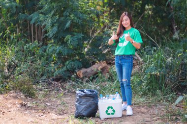 Güzel bir Asyalı kadın, dışarıda çöp kutusuna plastik şişe toplarken el işareti yapıyor.