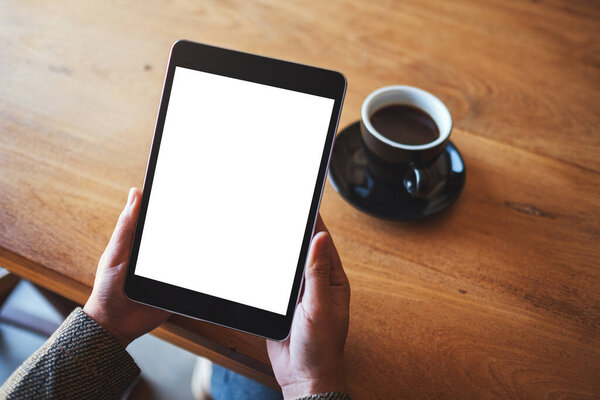Top view mockup image of hands holding black tablet pc with blank white screen with coffee cup on wooden table 