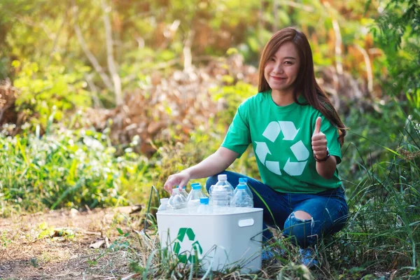 Güzel bir Asyalı kadın, dışarıda çöp kutusuna plastik şişe toplarken el işareti yapıyor.