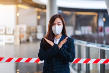 Korumacı yüz maskesi takan Asyalı bir kadının, Covid-19 'un yayılmasını önlemek için kırmızı ve beyaz uyarı bandı önünde çapraz kol işareti yaptığı yakın plan görüntüsü.