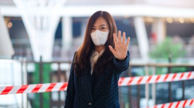 Korumacı yüz maskesi takan Asyalı bir kadının, Covid-19 'un yayılmasını önlemek için kırmızı ve beyaz uyarı bandının önünde dur işareti yaparken yakın plan görüntüsü.