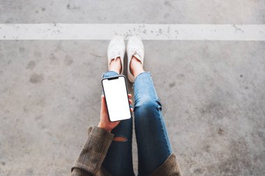 Üst görünümde, yerde otururken elinde beyaz ekranlı siyah bir cep telefonu tutan bir kadının görüntüsü var.