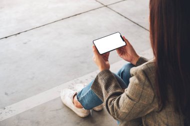 Yerde otururken elinde beyaz ekranlı siyah bir cep telefonu tutan bir kadının görüntüsü.