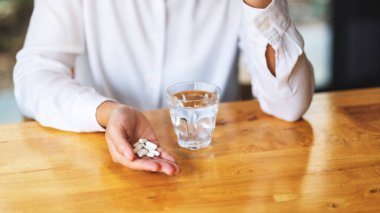 Closeup image of a woman holding white pills with a glass of water on the table
