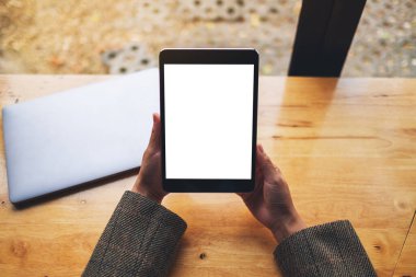 Masasında dizüstü bilgisayarı olan, boş beyaz masaüstü ekranlı, elinde siyah tablet bilgisayar tutan bir kadının en üstteki görünüm görüntüsü 