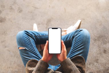 Üst görünümde, yerde otururken elinde beyaz ekranlı siyah bir cep telefonu tutan bir kadının görüntüsü var.