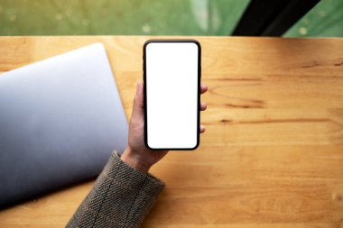 Masasında dizüstü bilgisayarı olan, boş beyaz masaüstü ekran cep telefonu tutan bir kadının en üstteki görüntüsü