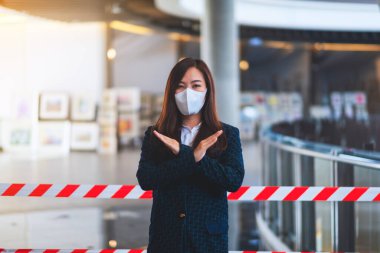 Korumacı yüz maskesi takan Asyalı bir kadının, Covid-19 'un yayılmasını önlemek için kırmızı ve beyaz uyarı bandı önünde çapraz kol işareti yaptığı yakın plan görüntüsü.