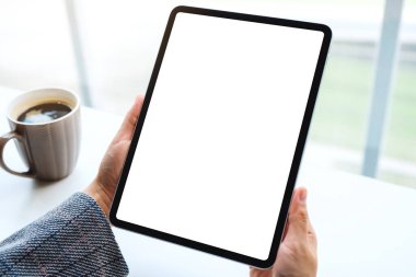 Masanın üzerinde kahve fincanı olan beyaz ekranlı siyah tableti tutan ellerin görüntüsü.